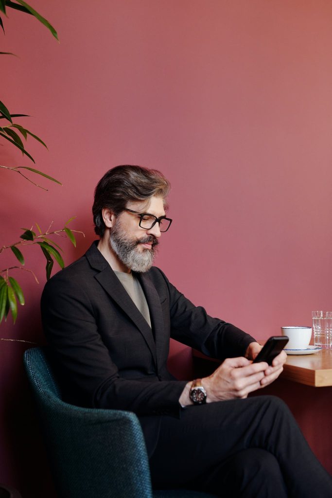 Photo of grey-bearded Man wearing Black Suit checking his phone.