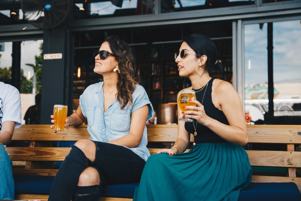 Woman in Black and Blue Sleeveless Dress Holding Glass of Beer Sitting on Bench.