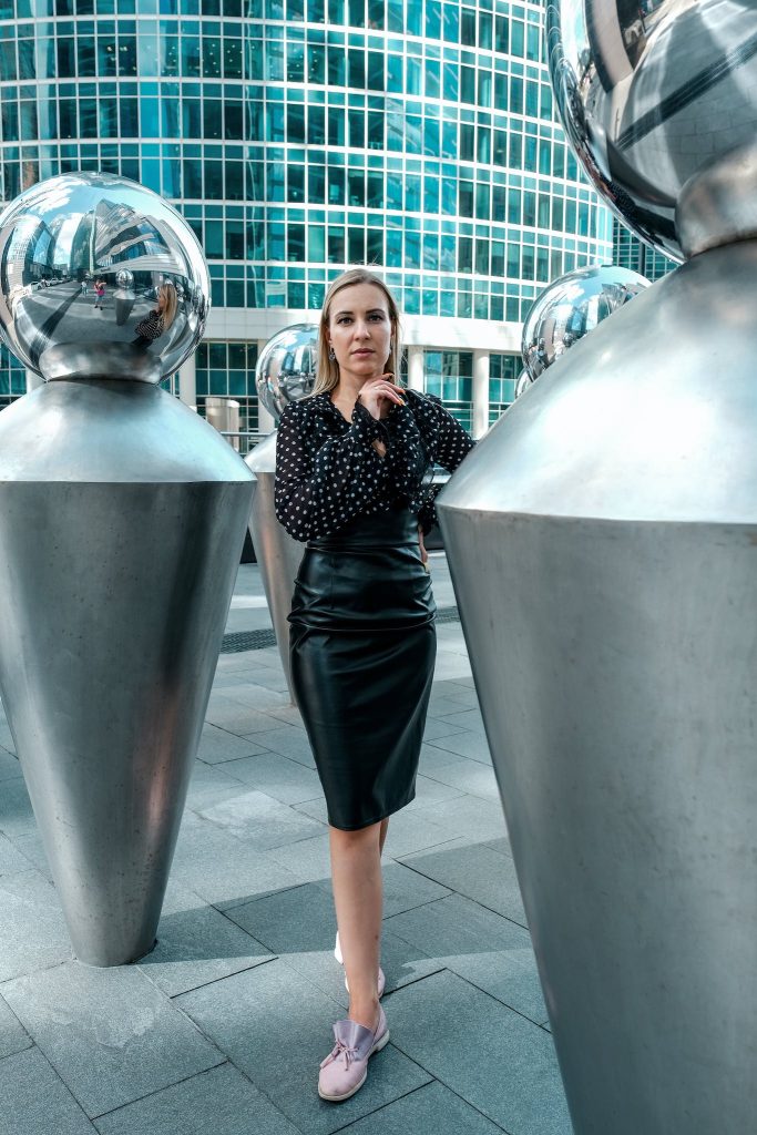 Full length independent female wearing trendy outfit touching chin in thoughts while standing amidst creative geometric urban decorations against modern skyscraper.